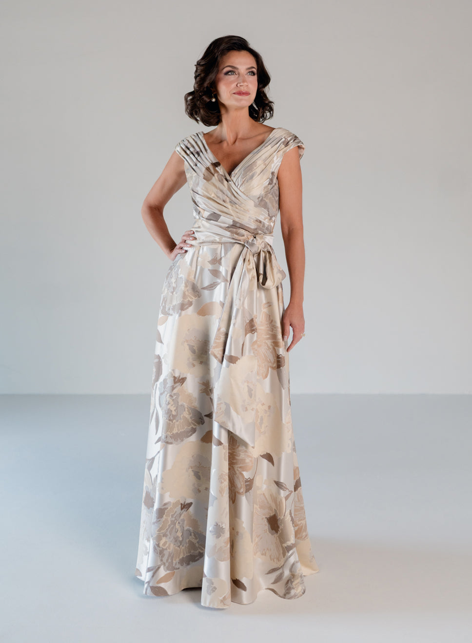 A woman modeling a floor-length wrap-style gown with a beige and taupe floral print, photographed against a plain light gray studio background. / A-line style in jacquard. Pleated front bodice with V-neck and trim with bow on left side front waist area.