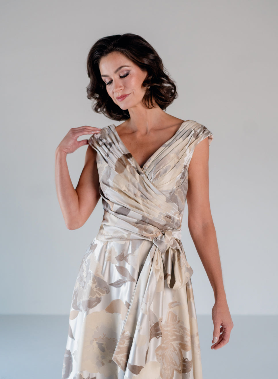 A woman modeling a sleeveless wrap-style dress with a tied waist, featuring a draped floral pattern in beige, taupe, and cream tones against a plain light gray studio background. / A-line style in jacquard. Pleated front bodice with V-neck and trim with bow on left side front waist area.