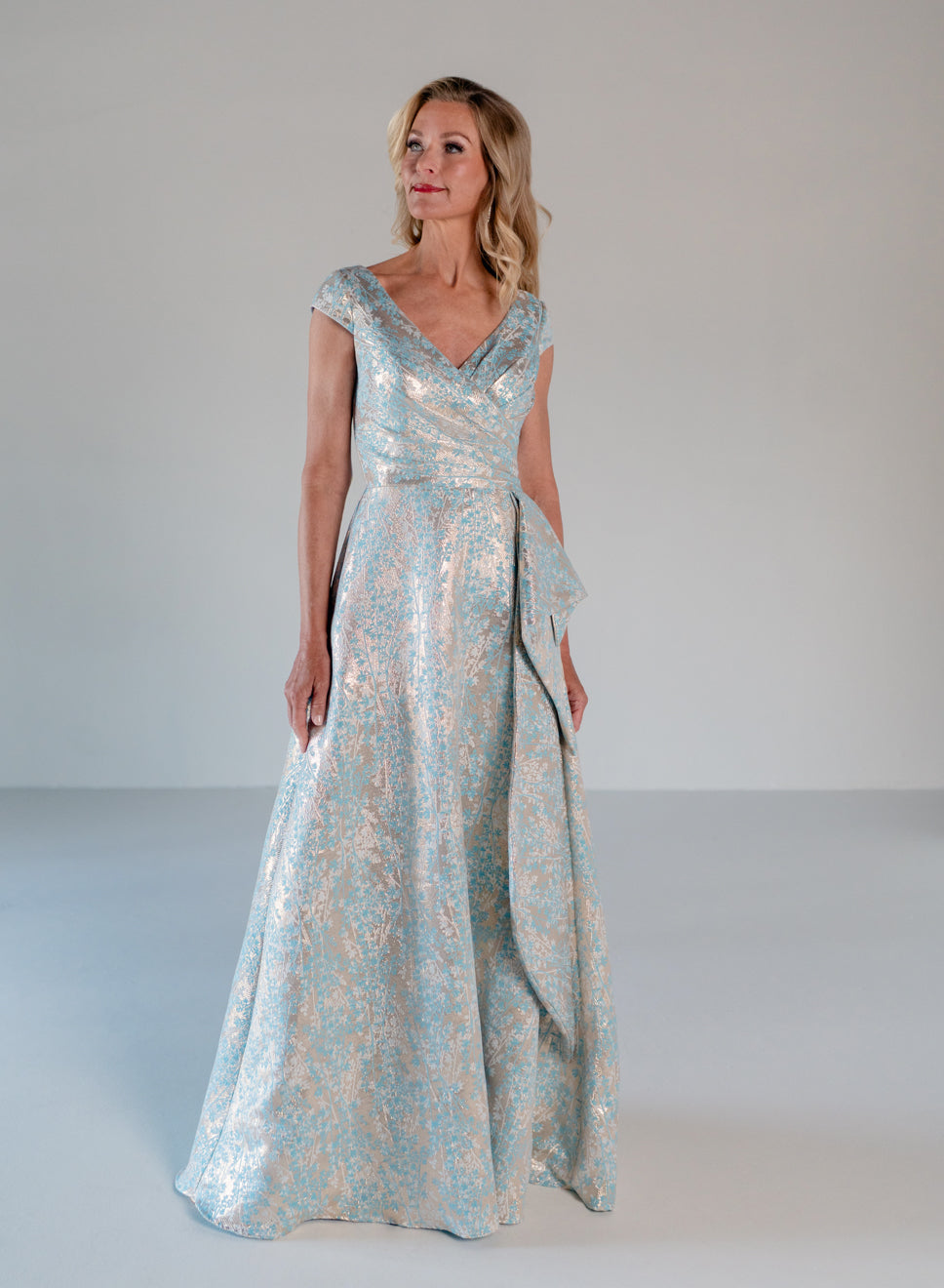 A woman modeling a floor-length formal gown with a shimmering light blue and gold floral pattern, standing against a neutral light gray studio background. / A-line style in soft brocade with V-neck and short cup sleeves, side draping on front bodice finished with draping panels on side front skirt.