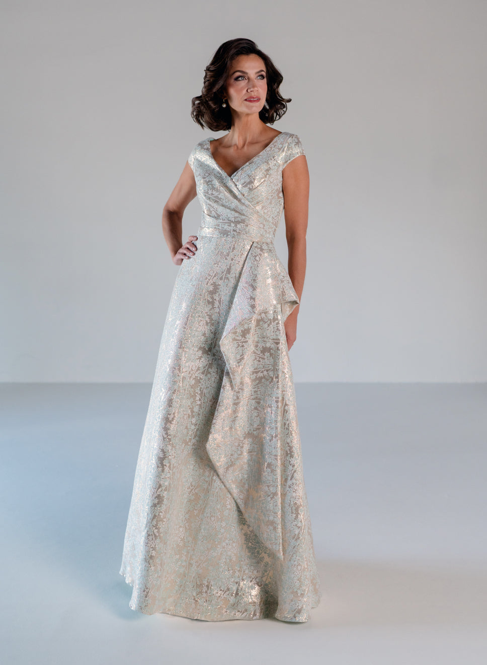 A woman modeling a floor-length formal gown with a shimmering silver and gold jacquard pattern, set against a plain light gray studio background. / A-line style in soft brocade with V-neck and short cup sleeves, side draping on front bodice finished with draping panels on side front skirt.
