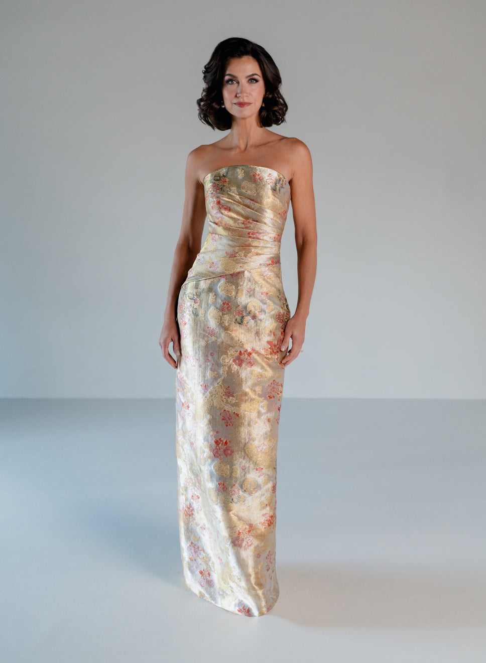 A woman modeling a strapless floor-length gown with a ruched bodice and a shimmering floral jacquard pattern in beige, gold, and muted pink, set against a plain light gray studio background. / Jacquard style with straight neckline and side draping on front bodice. Pencil skirt with split on CB.