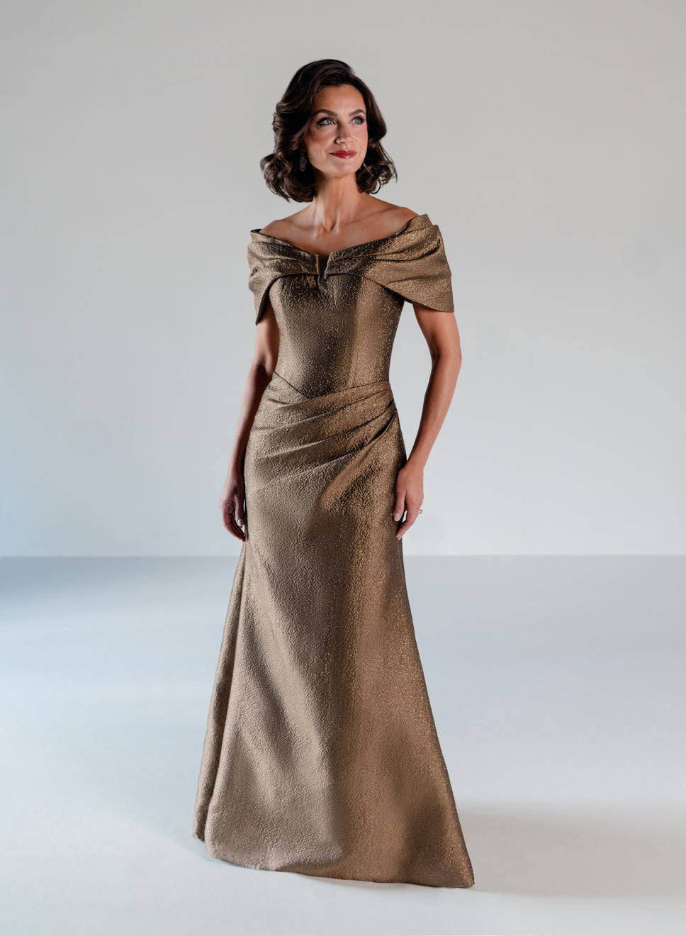 A woman modeling a floor-length off-the-shoulder gown in a shimmering metallic taupe-gold fabric against a plain light gray studio background. / A-line style in shimmer jacquard with off-shoulder collar and side front draping on skirt.