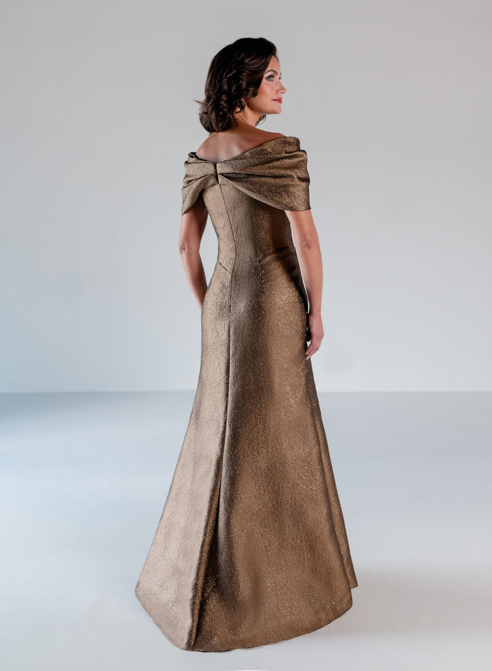 A woman modeling a floor-length metallic bronze gown with a draped off-the-shoulder neckline, shown from the back in a studio setting. / A-line style in shimmer jacquard with off-shoulder collar and side front draping on skirt.
