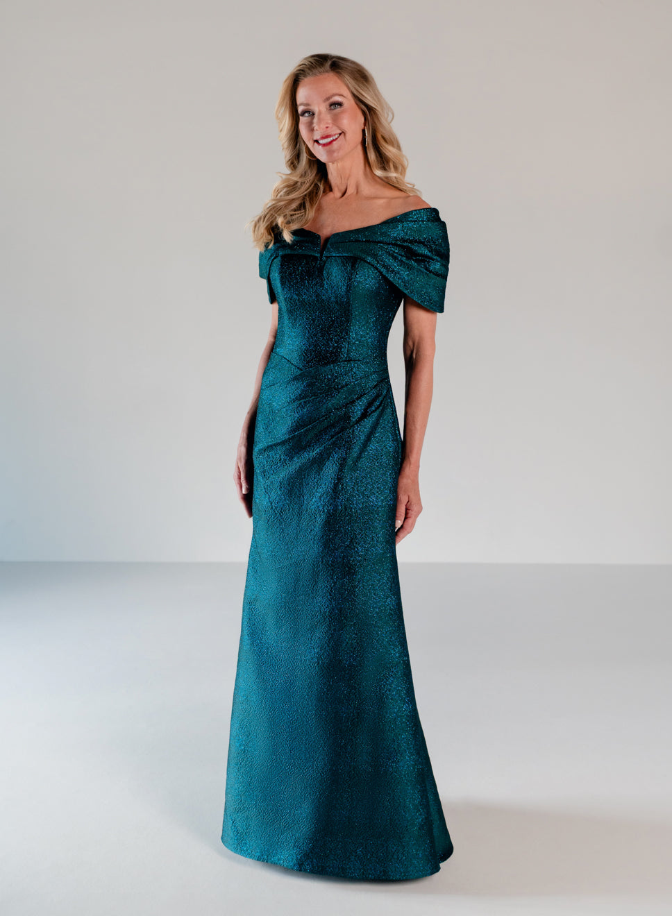 A woman modeling a floor-length off-the-shoulder gown in a rich teal green fabric with a subtle textured sheen, set against a plain light gray studio background. / A-line style in shimmer jacquard with off-shoulder collar and side front draping on skirt.