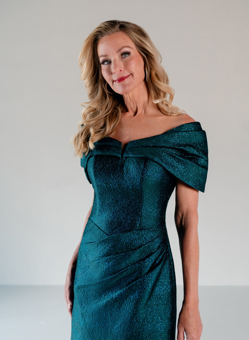 A woman modeling an off-the-shoulder teal green dress with a textured, shimmering finish against a neutral light gray background. / A-line style in shimmer jacquard with off-shoulder collar and side front draping on skirt.