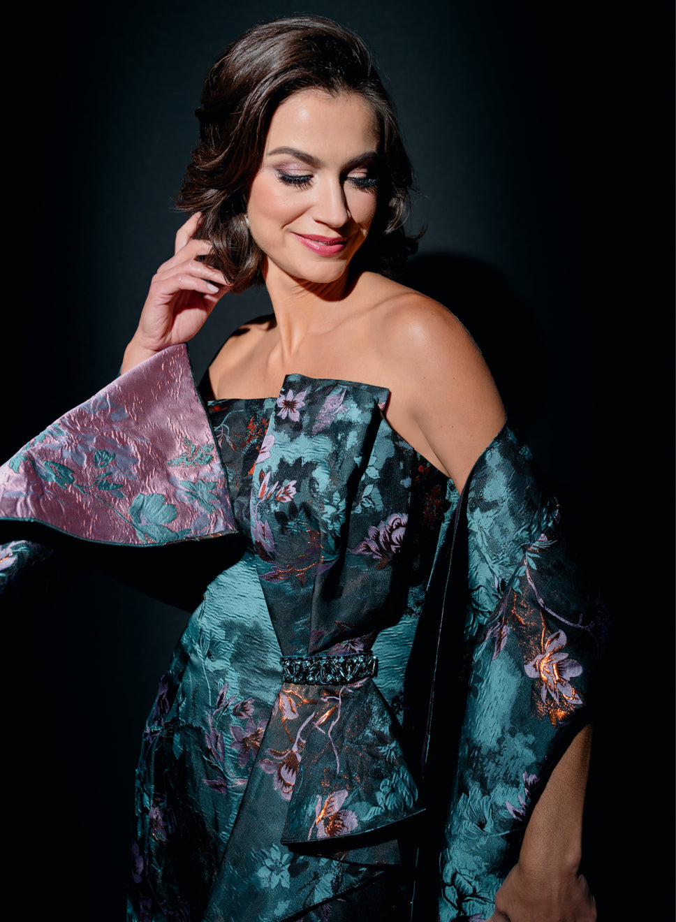 A woman wearing a strapless off-the-shoulder gown with a teal floral jacquard pattern and a matching draped capelet, posed against a dark studio background. / Slim style in jacquard with straight neckline and large bow on side front decorated with small, beaded detail.