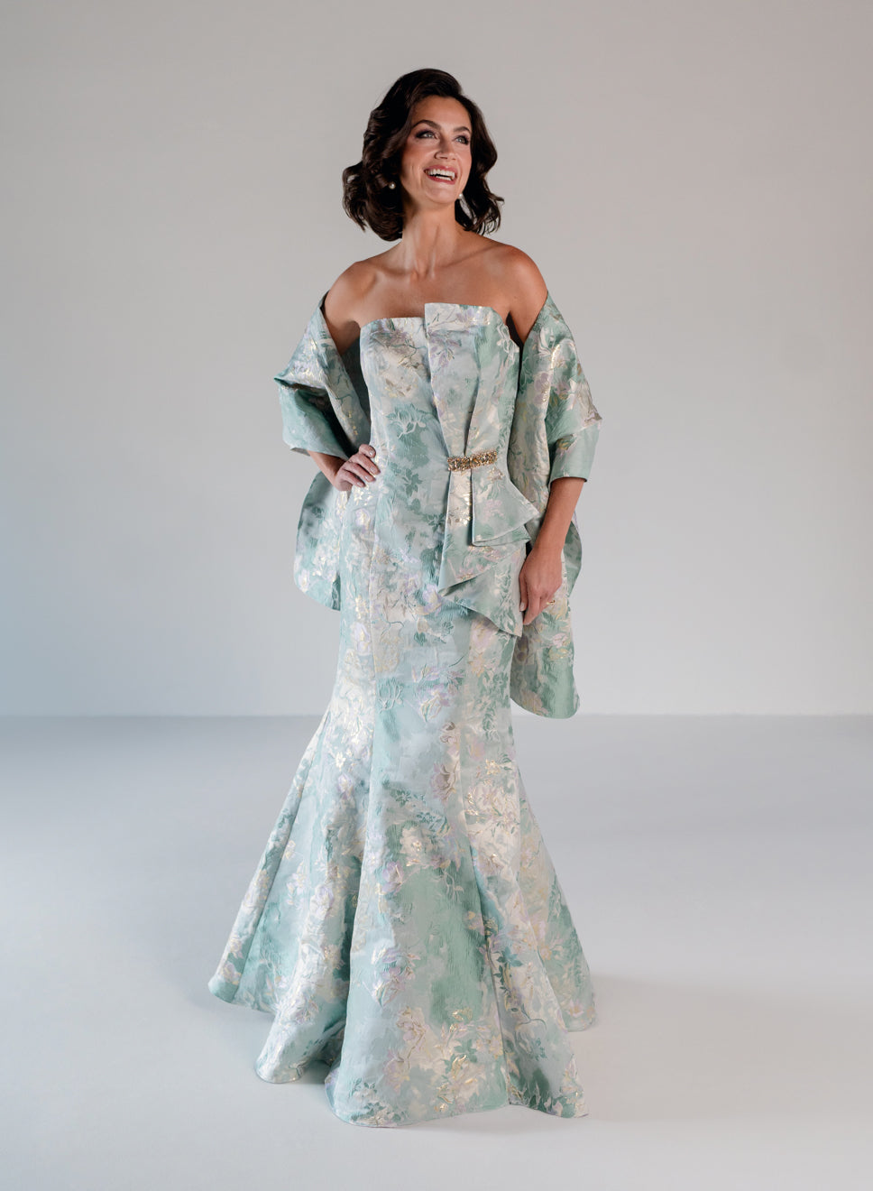 A woman modeling an off-the-shoulder formal gown with a matching draped shawl, featuring a light aqua and white floral jacquard pattern with a gold waist tie, set against a plain light gray studio background. / Slim style in jacquard with straight neckline and large bow on side front decorated with small, beaded detail.