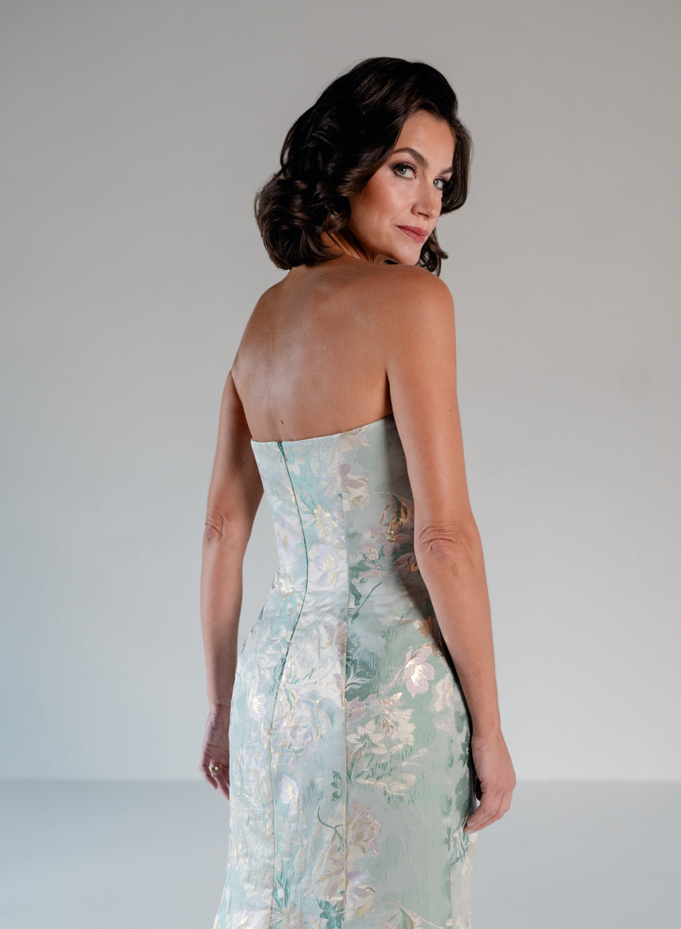 A woman wearing a strapless fitted gown with a pale mint green and white floral jacquard pattern, photographed from the back against a light gray studio background. / Slim style in jacquard with straight neckline and large bow on side front decorated with small, beaded detail.