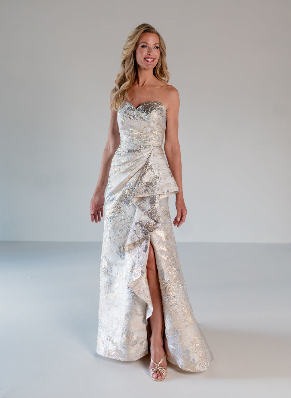 A woman modeling a strapless formal gown with a sweetheart neckline, ruched draped bodice, and a high-low ruffled hemline. The dress features a shimmering metallic silver and white jacquard pattern. / A-line brocade style with sweetheart neckline. Side draping on front dress finished with ruffle on side skirt area.