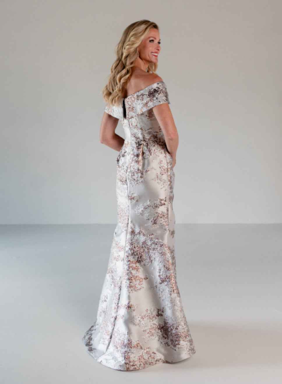 A woman modeling an off-the-shoulder floor-length gown with a metallic silver base and a rose-gold floral pattern, photographed in a studio setting. / A-line brocade style with sweetheart neckline. Side draping on front dress finished with ruffle on side skirt area.
