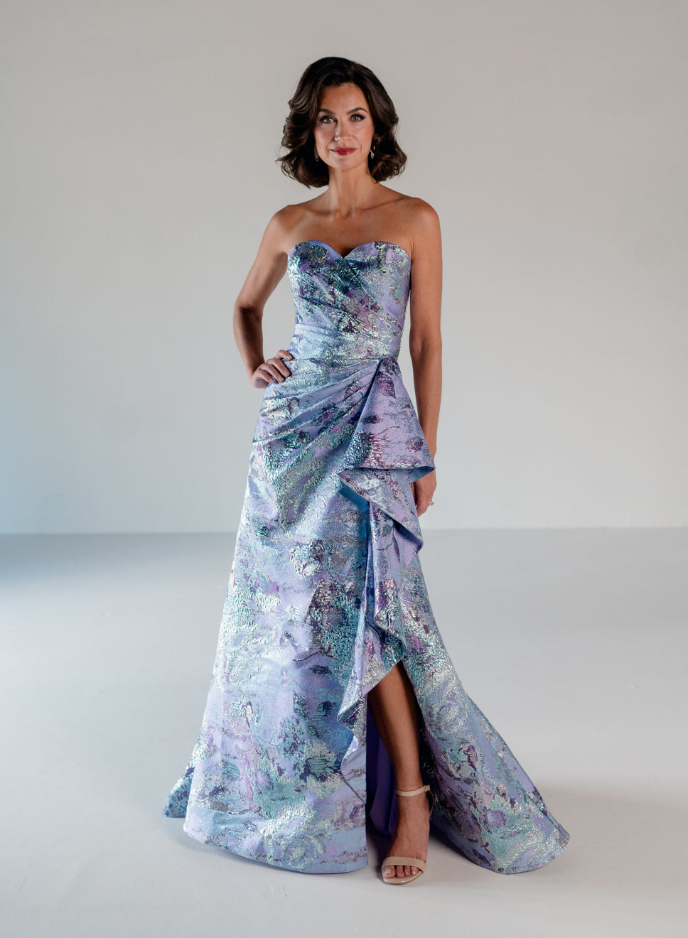 A woman models a strapless formal gown with a sweetheart neckline, ruched draped bodice, and a high-low skirt with a train. The dress features a shimmering abstract watercolor-like print in lavender, purple, blue, and green tones, set against a neutral light gray studio background. / A-line brocade style with sweetheart neckline. Side draping on front dress finished with ruffle on side skirt area.