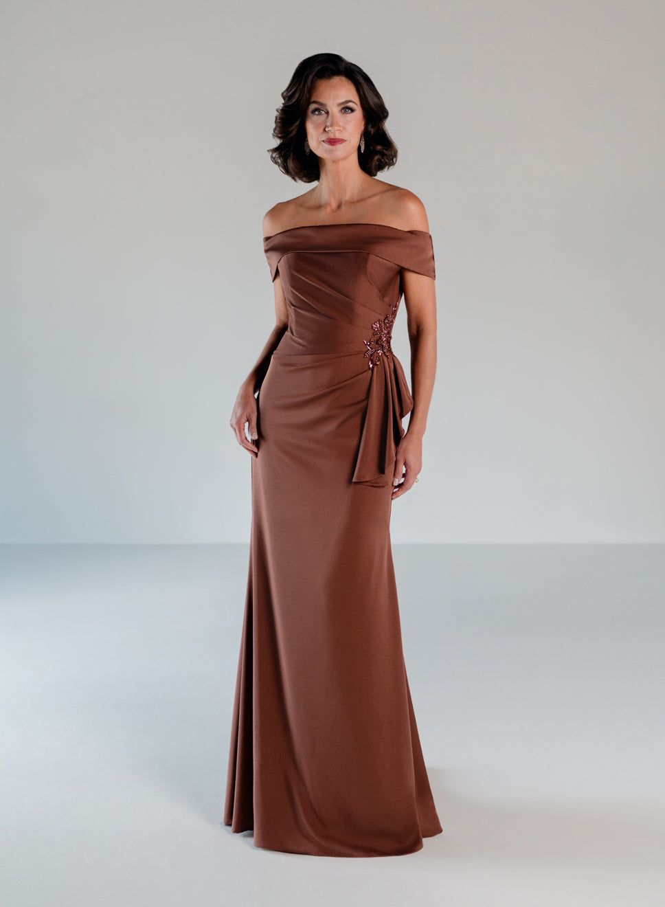 A woman modeling an off-the-shoulder floor-length gown in a warm brown shade, standing against a neutral light gray studio background. / A-line style in stretch satin with off-shoulder collar and side from draping finished with ruffle detail and beaded motif.