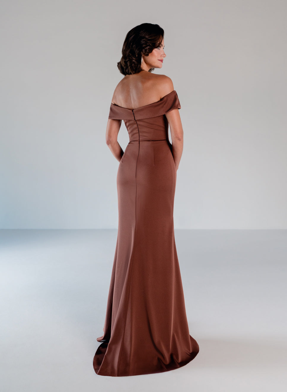 A woman modeling an off-the-shoulder floor-length gown in a warm brown or rust color, shown from the back in a studio setting with a light gray background. / A-line style in stretch satin with off-shoulder collar and side from draping finished with ruffle detail and beaded motif.