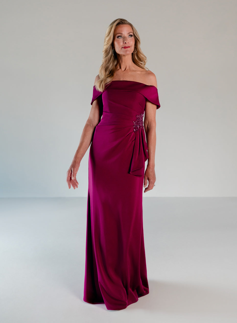 A woman modeling a floor-length off-the-shoulder gown in rich magenta-purple satin, featuring a draped waist detail and a small silver embellishment, set against a plain light gray studio background. / A-line style in stretch satin with off-shoulder collar and side from draping finished with ruffle detail and beaded motif.