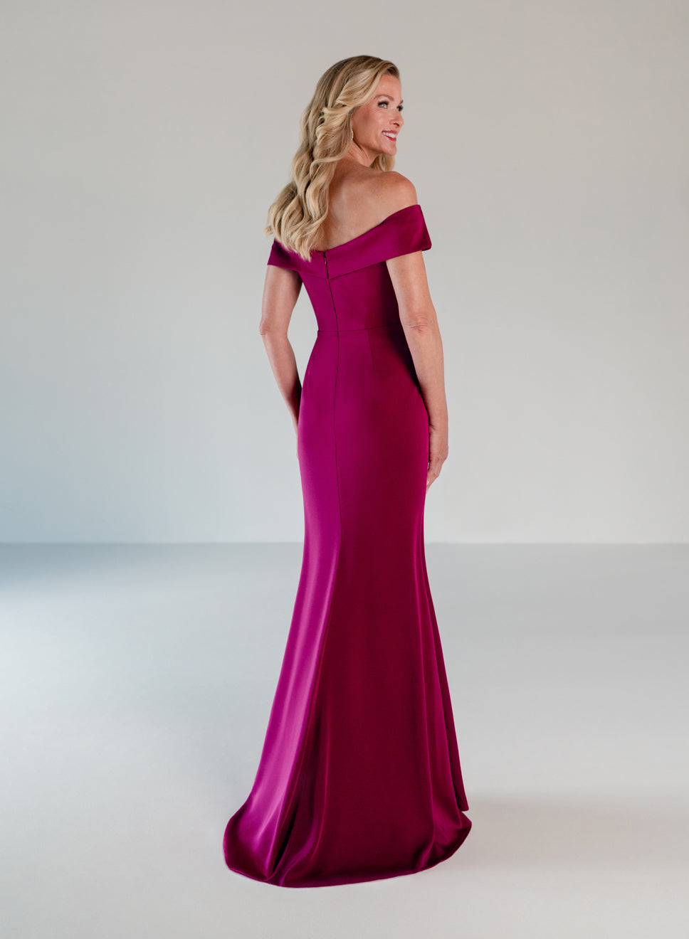 A woman modeling a floor-length off-the-shoulder evening gown in rich magenta satin, photographed from the back against a clean light gray studio backdrop. / A-line style in stretch satin with off-shoulder collar and side from draping finished with ruffle detail and beaded motif.