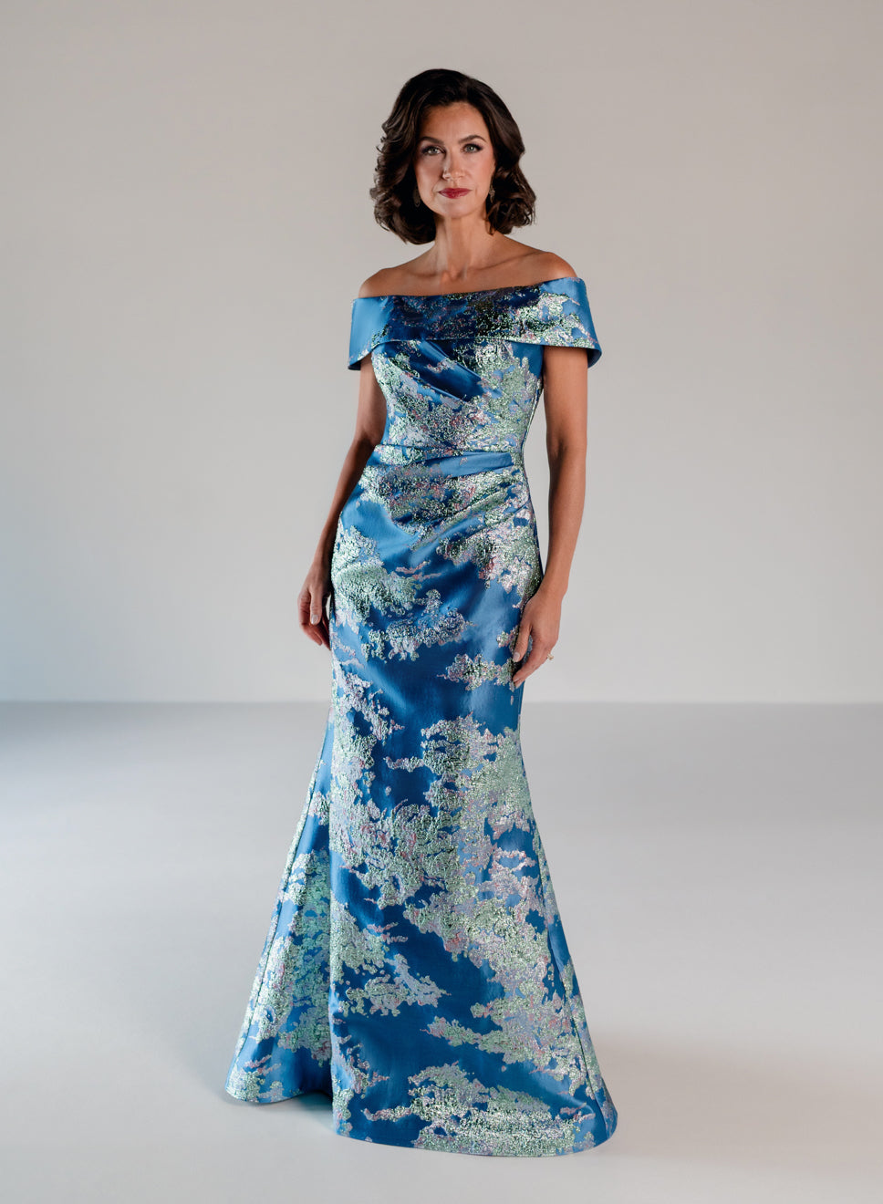 A woman modeling an off-the-shoulder blue satin gown with a light blue and white floral pattern, photographed against a neutral light gray studio background. / Slim style in brocade with straight neckline and off-shoulder collar. Side draping on front dress.