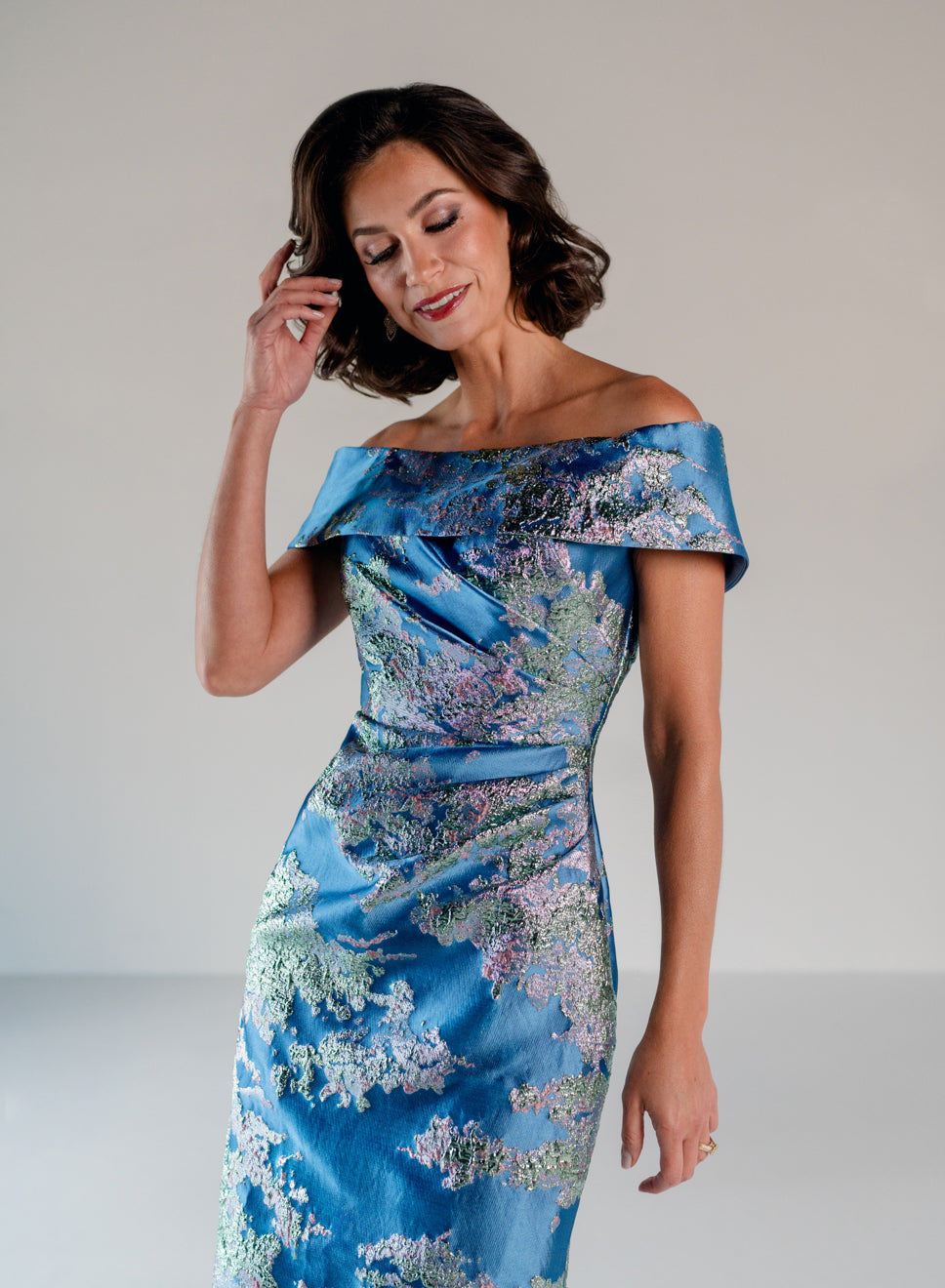 A woman modeling an off-the-shoulder blue satin dress with a floral jacquard pattern in shades of purple, green, and white, set against a plain light gray studio background. / Slim style in brocade with straight neckline and off-shoulder collar. Side draping on front dress.
