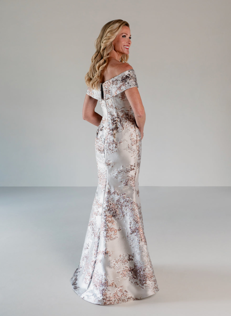 A woman modeling an off-the-shoulder floor-length gown with a shimmering silver base and a rose-gold floral pattern, photographed against a neutral light gray studio background. / Slim style in brocade with straight neckline and off-shoulder collar. Side draping on front dress.