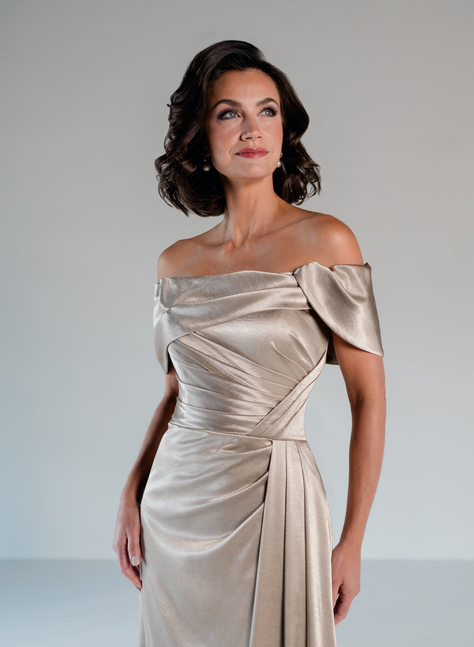 A woman modeling an off-the-shoulder champagne satin gown with draped detailing and a fitted waist, photographed against a neutral light gray studio background. / Slim A-line style in smooth charmeuse with off-shoulder collar, asymmetrical pleating on front dress.