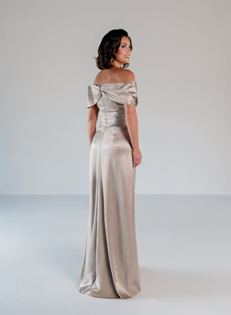 A woman modeling an off-the-shoulder floor-length satin gown in a champagne beige color, photographed against a neutral light gray studio background. / Slim A-line style in smooth charmeuse with off-shoulder collar, asymmetrical pleating on front dress.