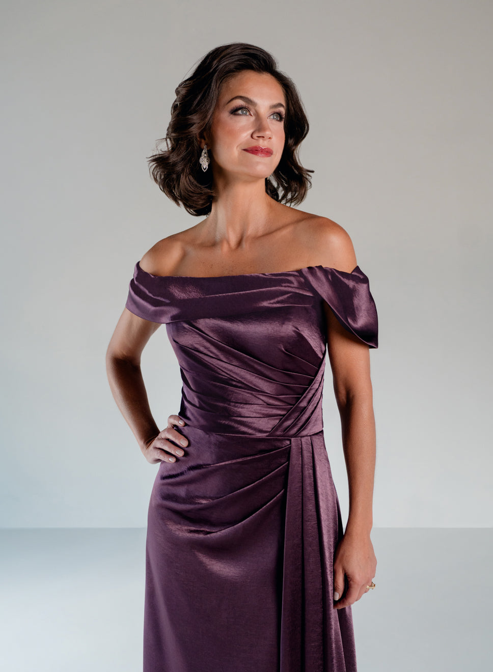 A woman modeling an off-the-shoulder, floor-length purple satin dress with draped detailing and a gathered waist, photographed against a light gray studio background. / Slim A-line style in smooth charmeuse with off-shoulder collar, asymmetrical pleating on front dress.