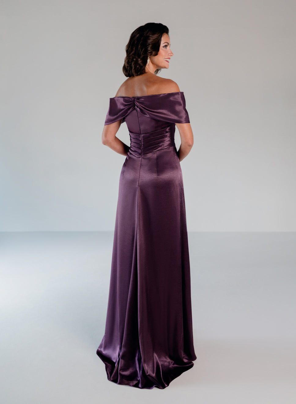 A woman modeling a floor-length off-the-shoulder gown in a deep plum purple satin fabric, shown from the back against a neutral light gray studio background. / Slim A-line style in smooth charmeuse with off-shoulder collar, asymmetrical pleating on front dress.