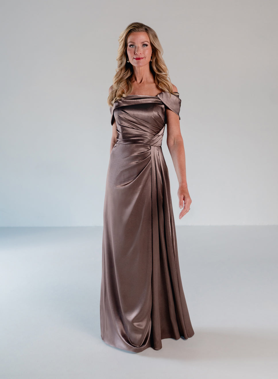 A woman modeling a floor-length off-the-shoulder gown in a satin-like taupe-brown fabric against a light gray studio background. / Slim A-line style in smooth charmeuse with off-shoulder collar, asymmetrical pleating on front dress.