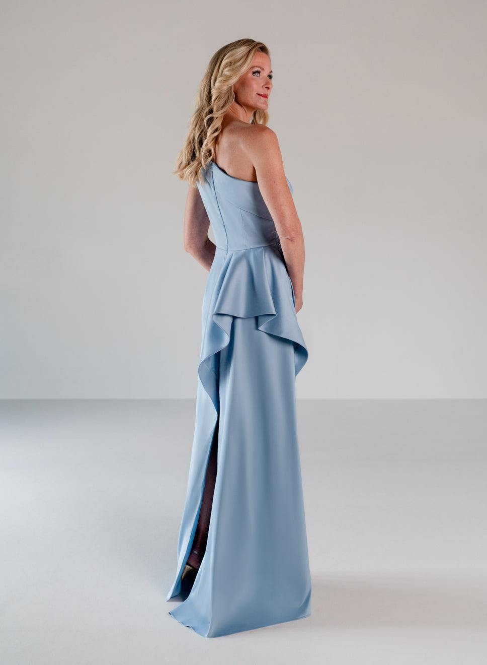 A woman modeling a light blue one-shoulder evening gown with a peplum waist and high side slit, photographed in a minimalist studio setting. / Draped column gown with placed beading.