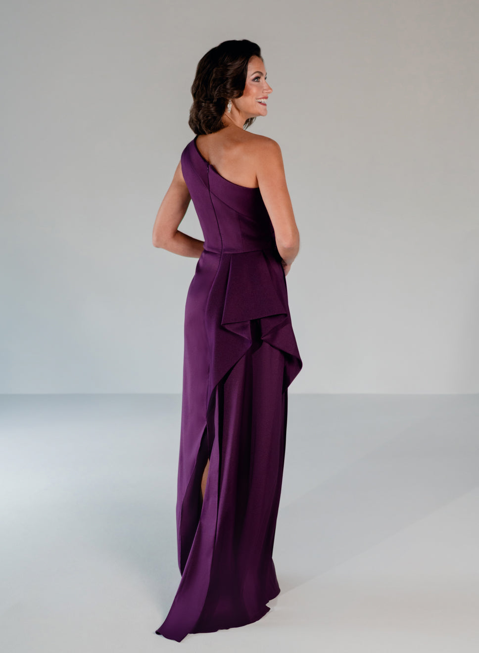 A woman modeling a floor-length one-shoulder gown in deep purple, shown from the back and side against a plain light gray studio background. / Draped column gown with placed beading.