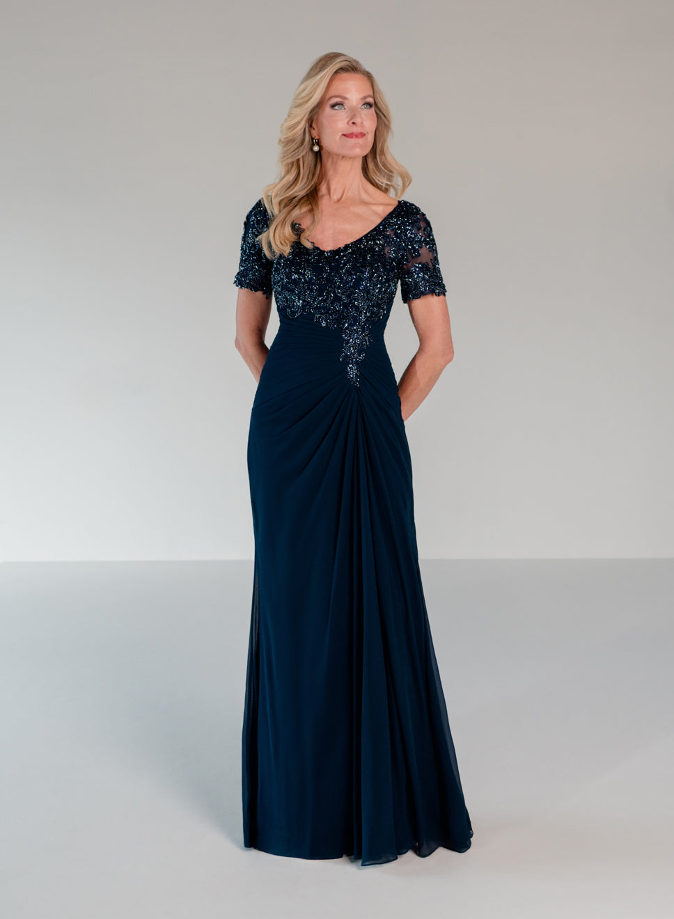 A woman modeling a floor-length navy evening gown with a beaded lace bodice, short sleeves, and a draped skirt, photographed against a neutral light gray studio background. / Slim A-line style with chiffon skirt and beaded lace bodice, V-neck with short sleeves and side front draping on skirt.