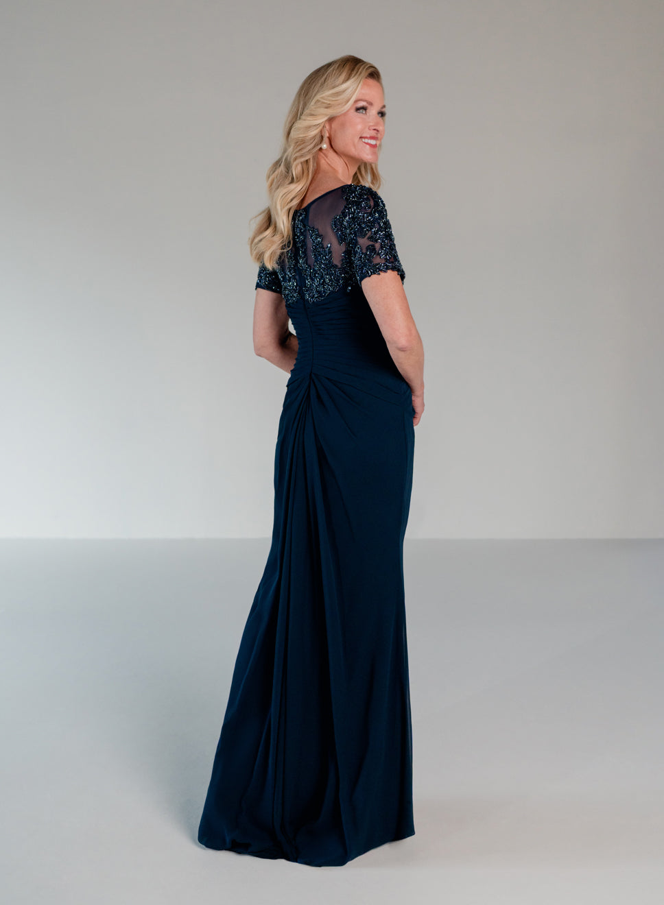 A woman modeling a floor-length navy blue evening gown from the back in a studio setting with a light gray background. / Slim A-line style with chiffon skirt and beaded lace bodice, V-neck with short sleeves and side front draping on skirt.