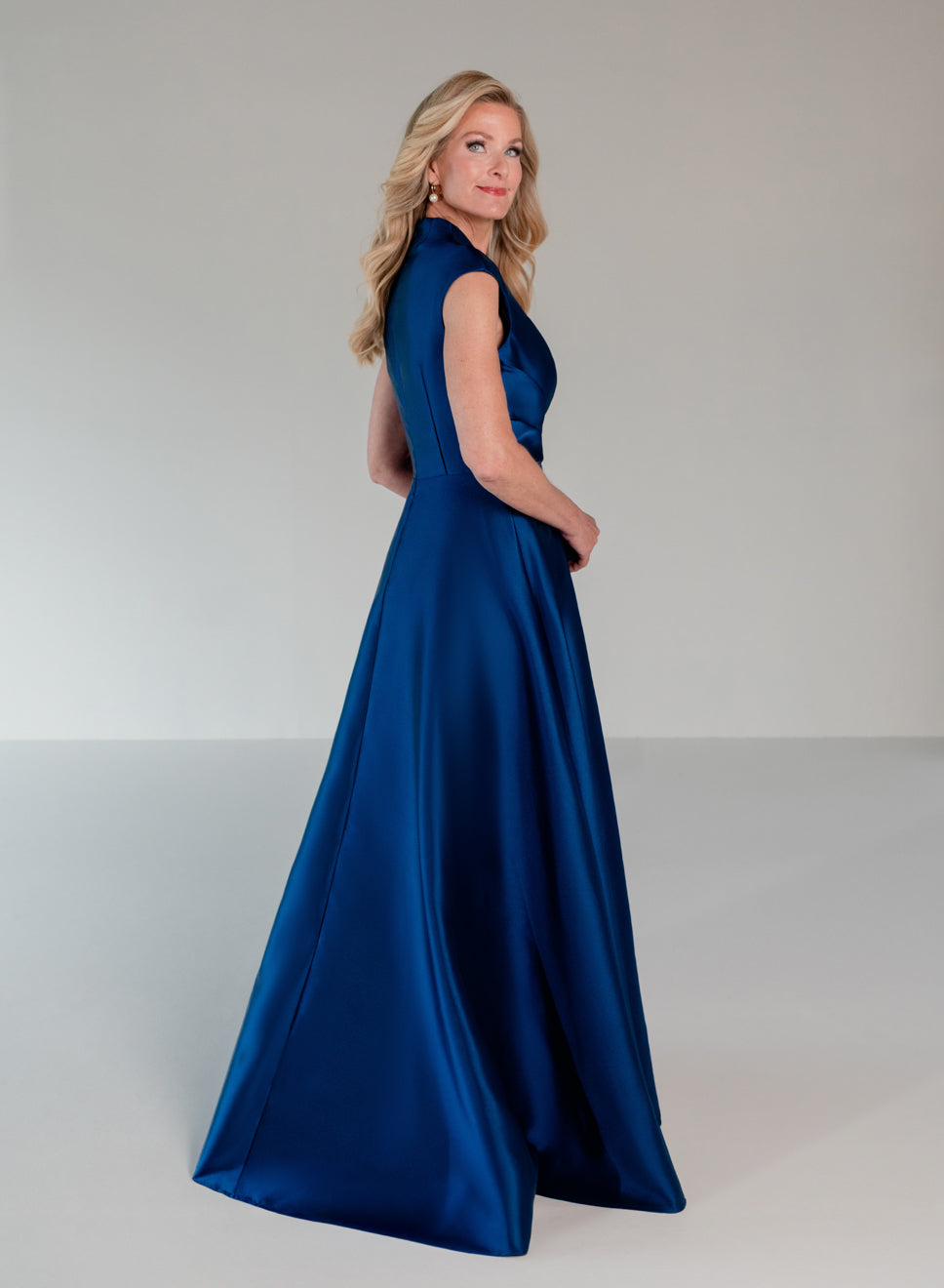 A woman modeling a floor-length royal blue satin gown with a sleeveless V-neckline and structured back, photographed in a clean studio setting. / A-line mikado style with V-neck and short cup sleeves. Side front draping on bodice finished with ruffle on side front skirt.
