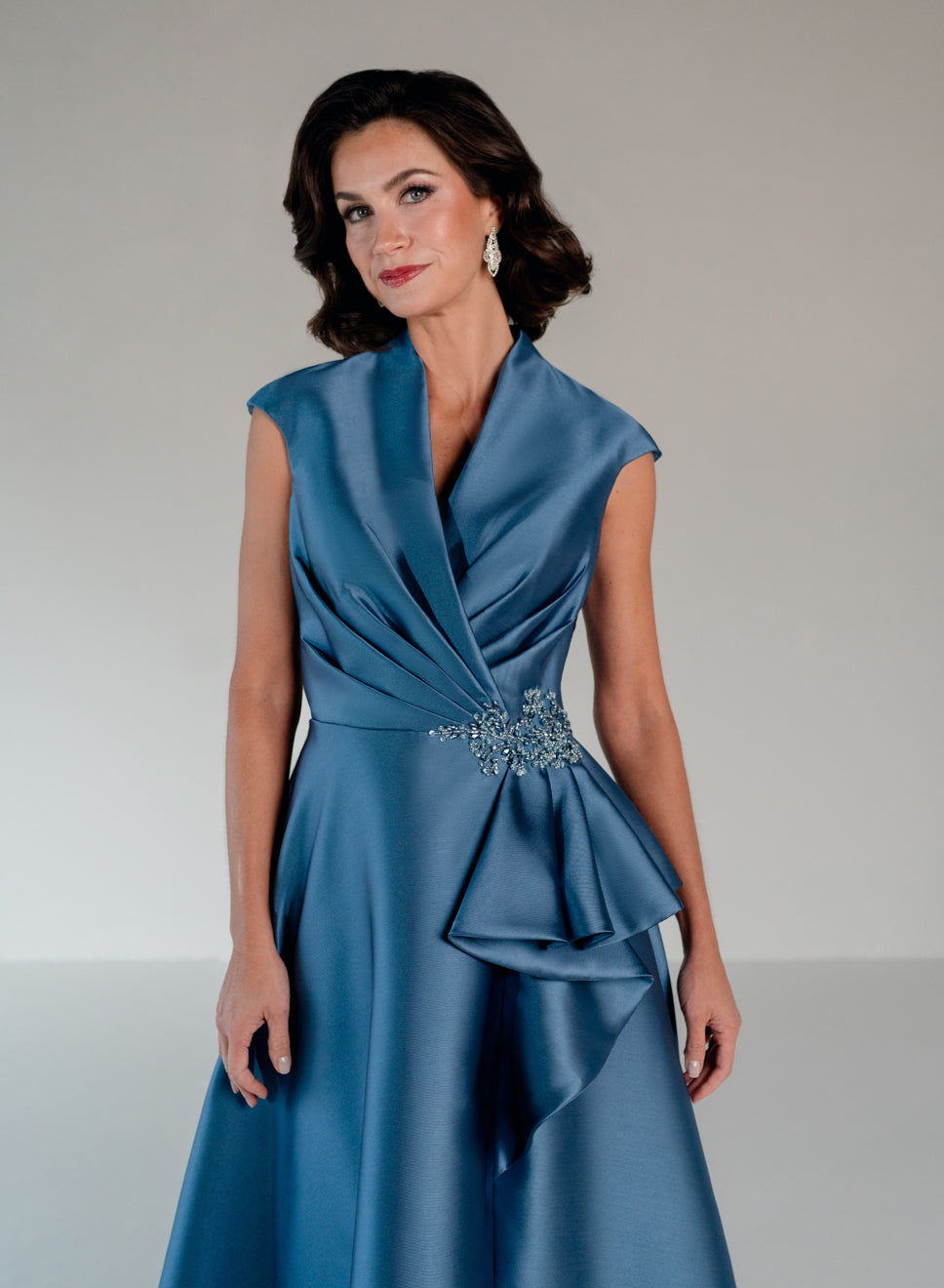 A woman modeling a sleeveless blue satin dress with a draped wrap-style bodice, peplum-like ruffle at the waist, and a decorative jeweled embellishment at the waist, against a plain light gray studio background. / A-line mikado style with V-neck and short cup sleeves. Side front draping on bodice finished with ruffle on side front skirt.