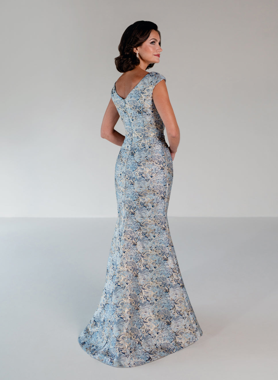 A woman modeling a floor-length, fitted evening gown with a floral lace pattern in light blue, beige, and white, photographed against a plain light gray studio background. / Slim brocade style with soft V-neck and small cup sleeves. Side draping on front dress.