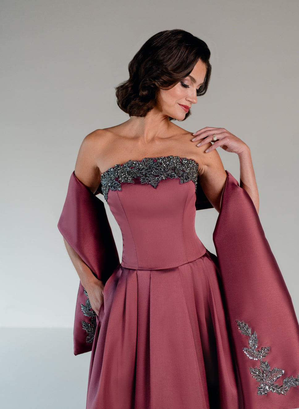 A woman wearing a strapless dusty rose evening gown with a structured corset bodice, a full skirt, and draped cape sleeves with silver beaded embellishments, posed against a neutral gray studio background. / Mikado A-line ballgown with dropped waist and placed beading at the upward scoop neckline.