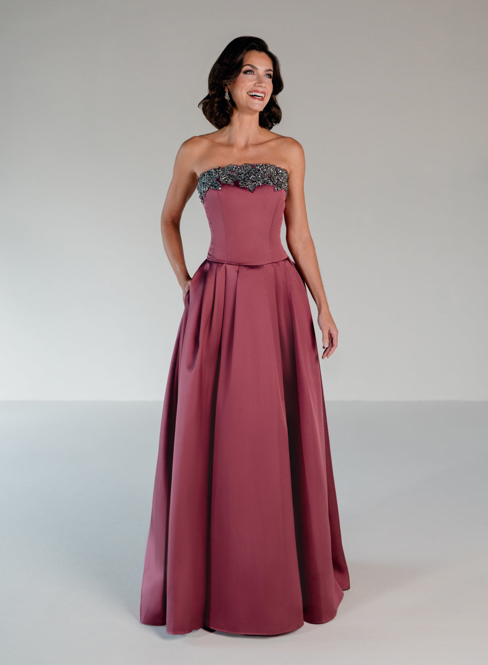 A woman modeling a strapless floor-length gown in dusty rose with a dark beaded lace bodice, photographed against a neutral light gray studio background. / Mikado A-line ballgown with dropped waist and placed beading at the upward scoop neckline.