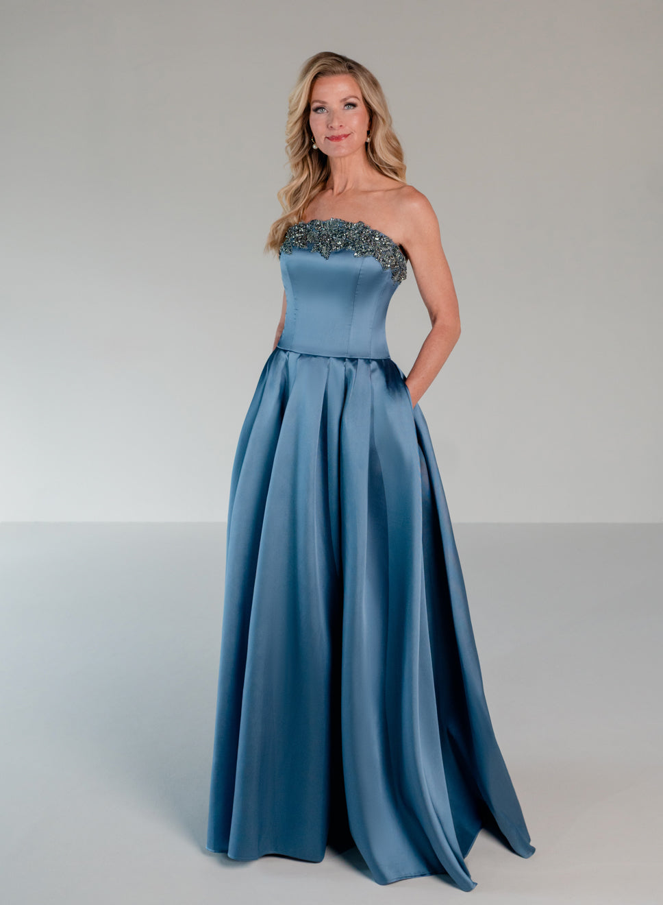 A woman modeling a floor-length formal gown in a dusty blue satin fabric with a beaded strapless bodice, standing against a neutral light gray studio background. | Mikado A-line ballgown with dropped waist and placed beading at the upward scoop neckline.