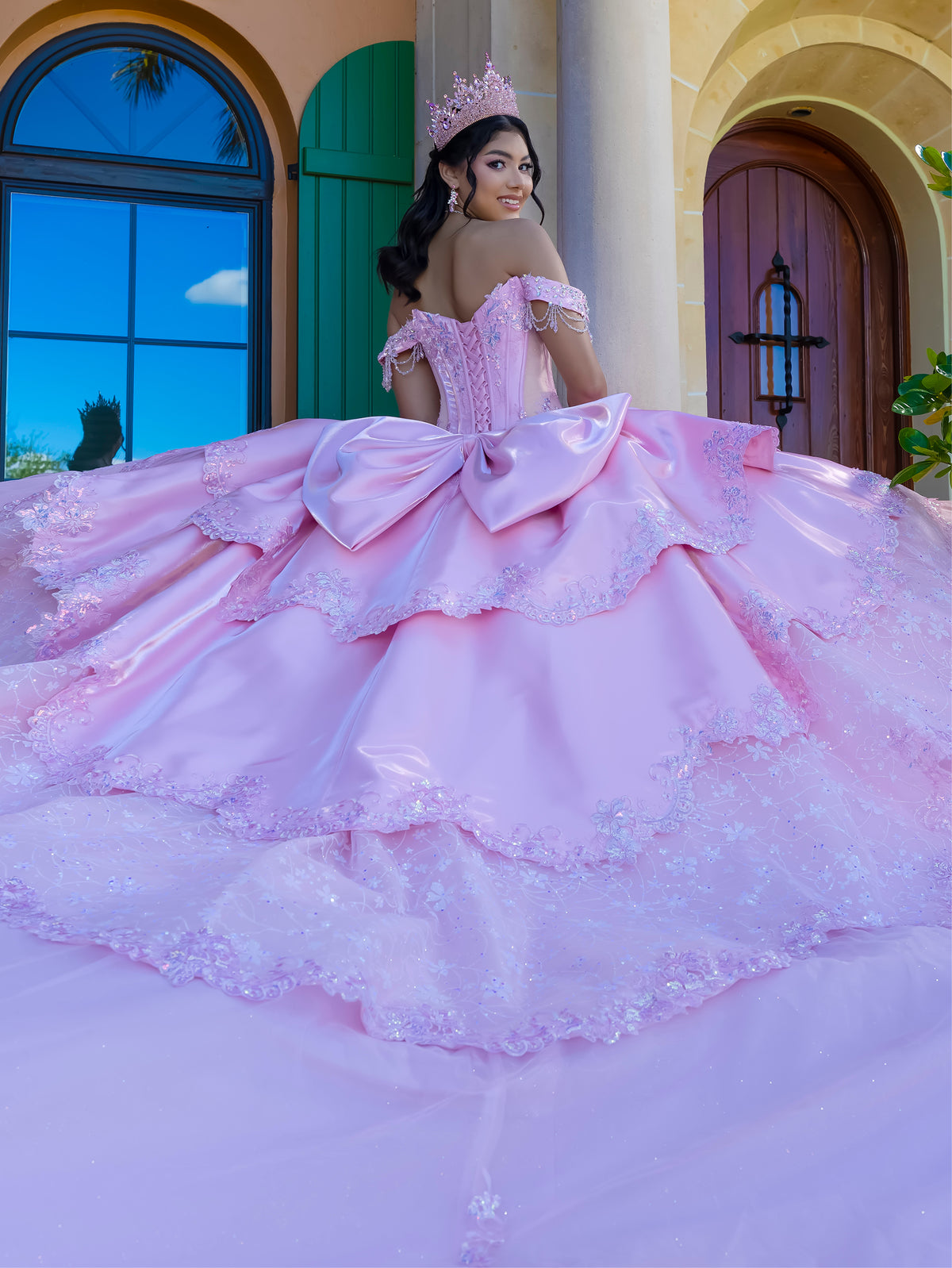 A woman wearing a voluminous light pink ball gown with a large bow at the back, off-the-shoulder lace detailing, and a matching tiara, posed in front of a warm-toned building with arched windows and doors. / This gown features a fitted, embellished bodice with off-the-shoulder sleeves and a full tiered ball-gown skirt. Intricate appliqués decorate the layers, and a long flowing train with a large back bow completes the princess-style look.