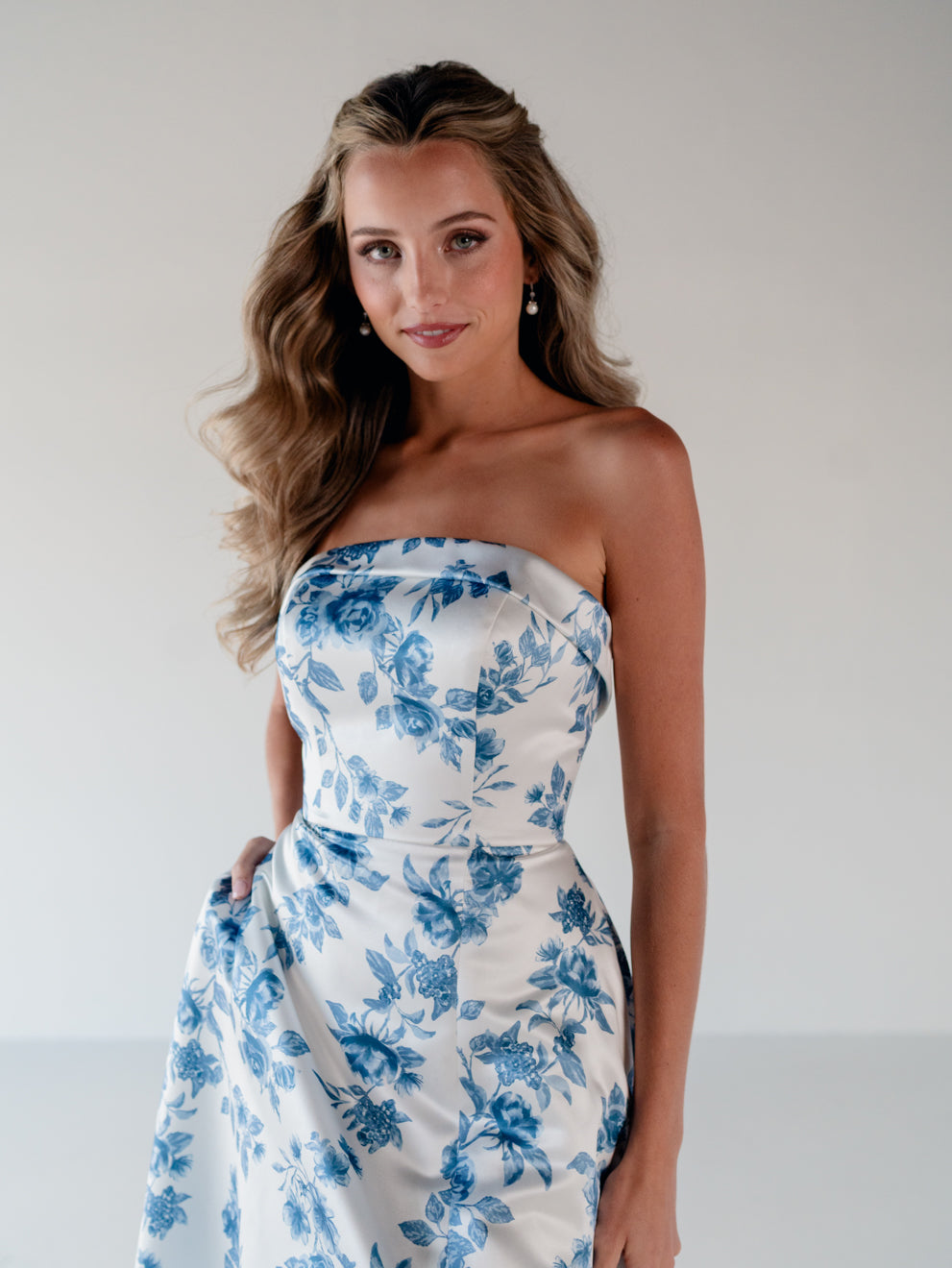 A woman modeling a strapless white dress with a blue floral toile pattern, standing against a plain light gray studio background. / Straight neckline A-line gown with back bow detail.