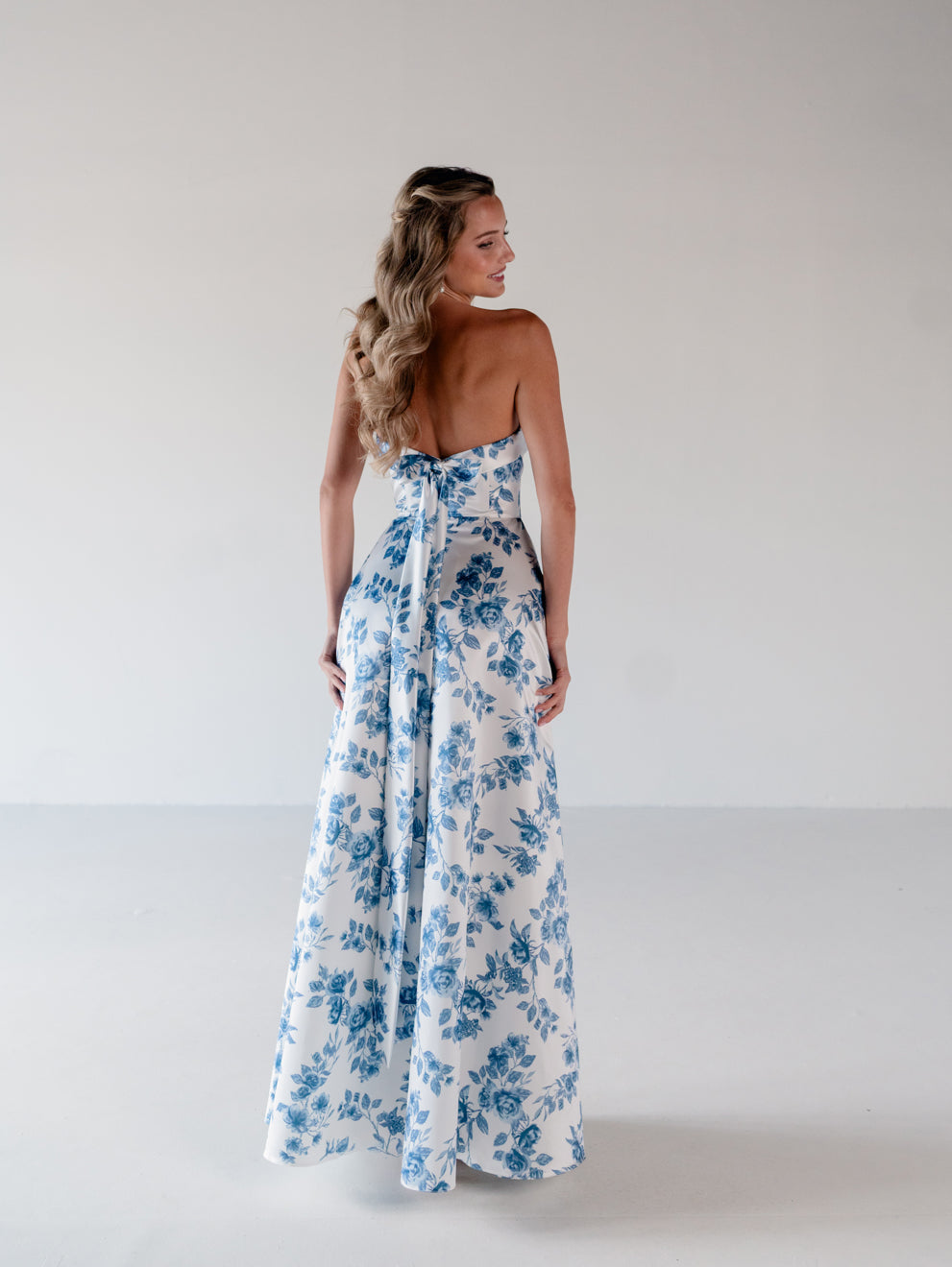 A woman stands with her back to the camera wearing a strapless white maxi dress with a blue floral print and a tie detail at the back, set against a plain light gray studio background. / Straight neckline A-line gown with back bow detail.