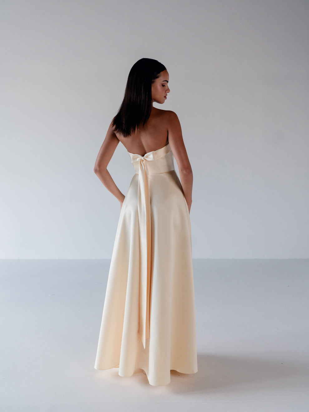 A woman stands with her back to the camera wearing a strapless cream satin gown with a large bow at the back of the neckline and a flowing floor-length skirt, photographed in a minimalist studio setting. / Straight neckline A-line gown with back bow detail.