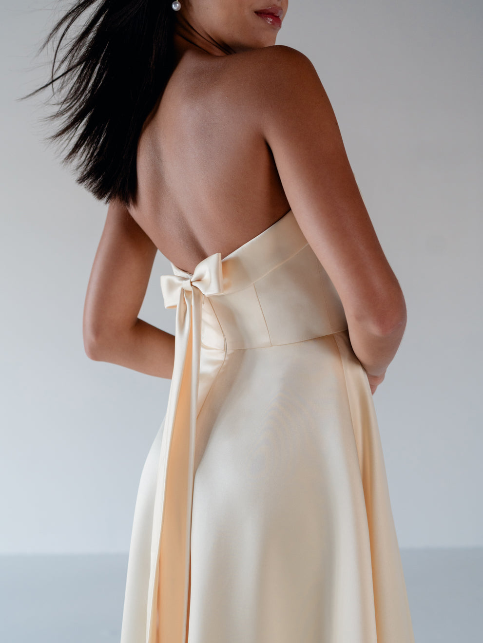 A woman shown from the back wearing a strapless satin dress with a large bow detail at the back, in a soft cream or pale peach tone against a light gray studio background. / Straight neckline A-line gown with back bow detail.