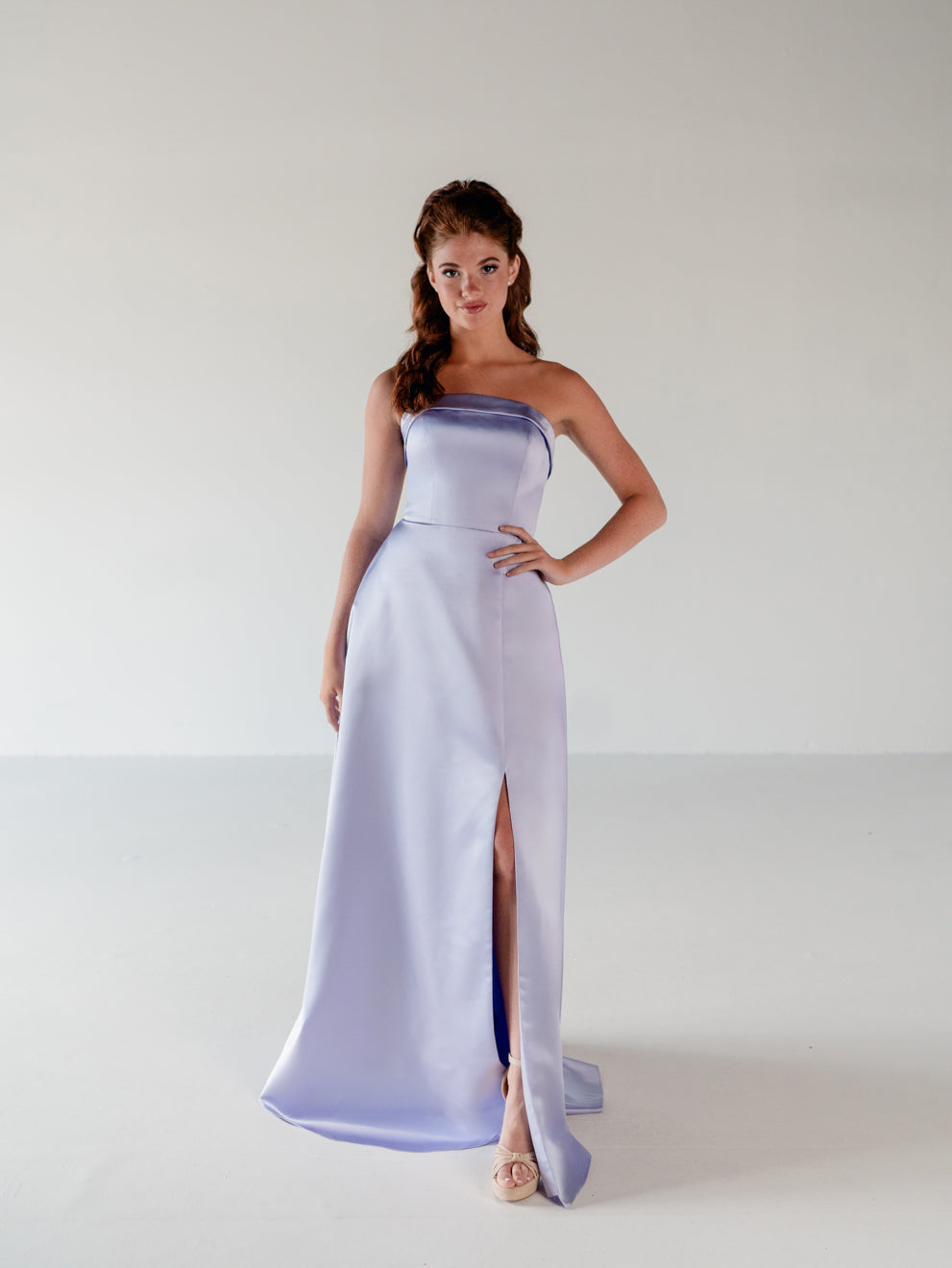 A woman modeling a strapless lavender satin maxi gown with a thigh-high slit, standing against a plain light gray studio background. / Straight neckline A-line gown with back bow detail.
