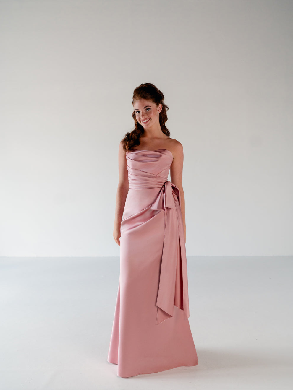 A young woman modeling a strapless dusty pink satin maxi dress with a draped waist and side bow, set against a plain light gray studio background. / Slim column gown with draped bodice and bow detail.