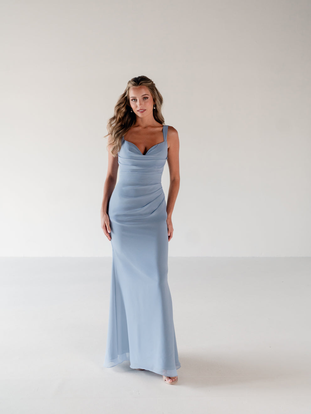 A woman modeling a floor-length light blue sleeveless gown with a draped, ruched bodice and a fitted skirt, standing against a plain light gray studio background. / Slim chiffon gown with bodice and skirt pleats and a cowl back.