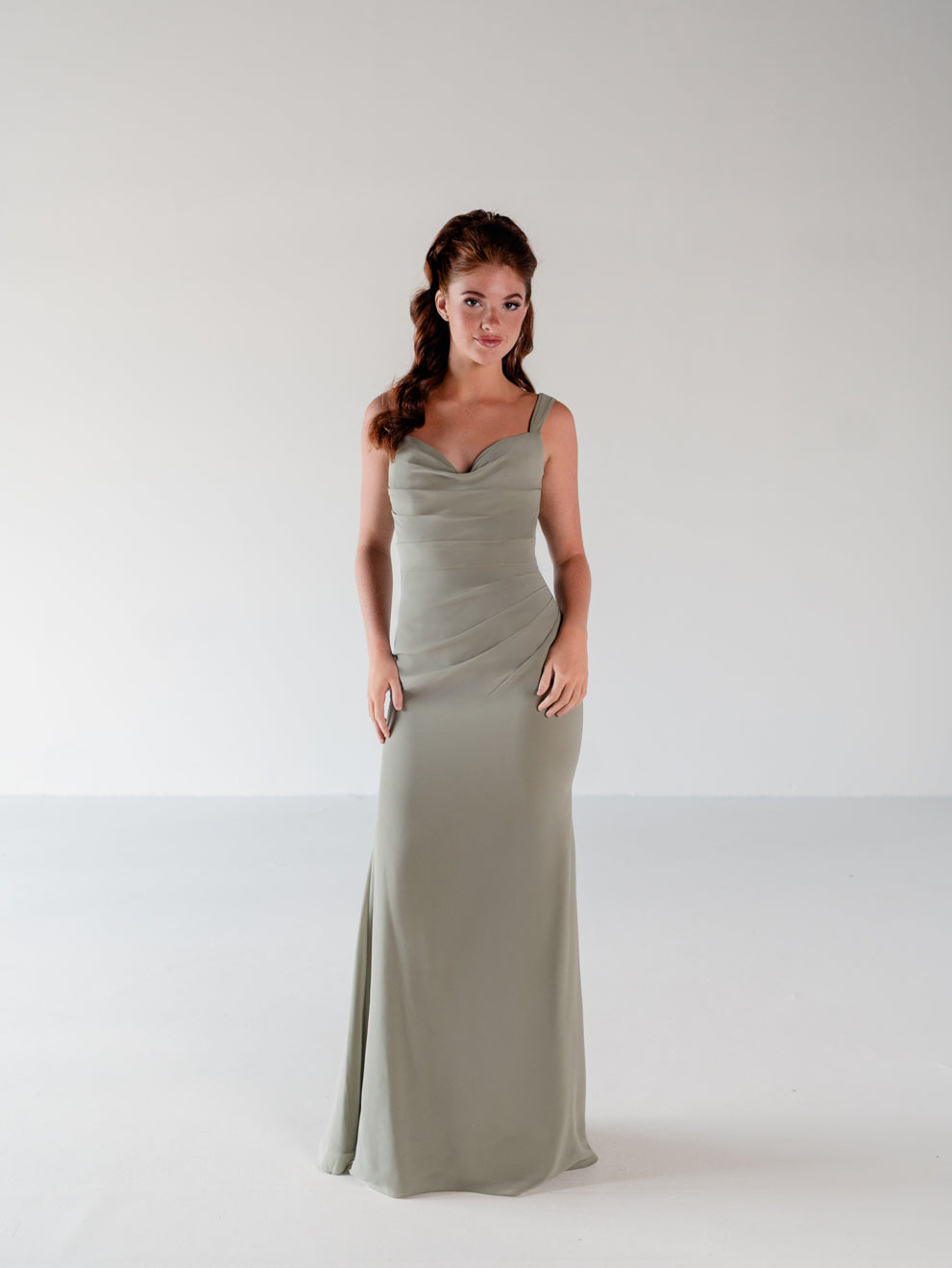 A woman modeling a floor-length, sleeveless cowl-neck dress in a muted sage green color, standing against a seamless light gray studio background. / Slim chiffon gown with bodice and skirt pleats and a cowl back.