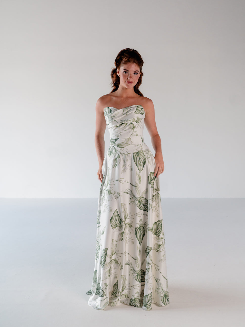 A woman modeling a strapless white maxi dress with a green leaf pattern, standing against a plain light gray studio background. / Basque waist draped bodice with an A-line skirt.