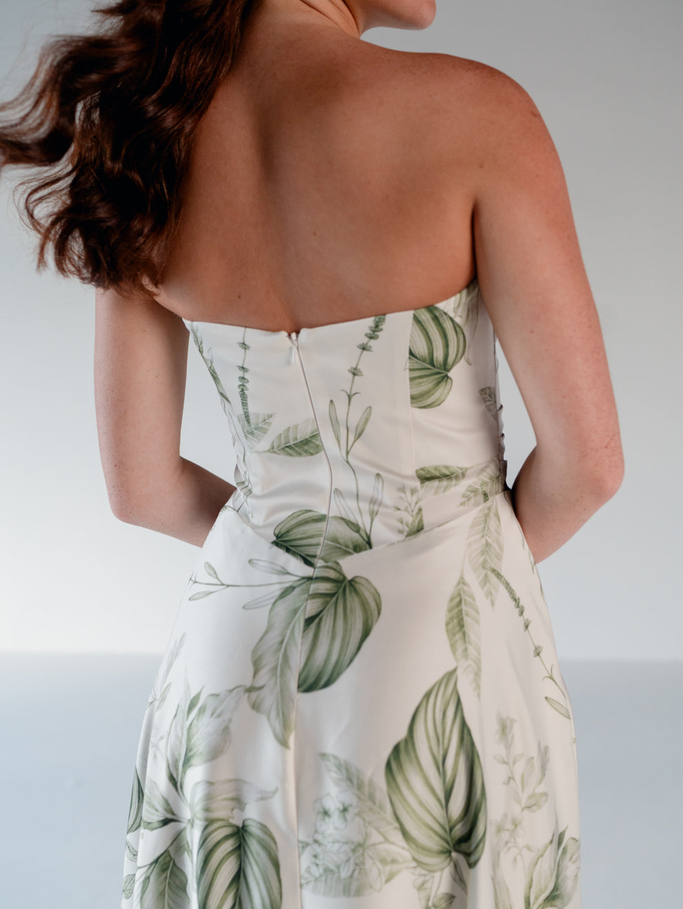 A woman shown from the back wearing a strapless dress with a green botanical leaf print on a warm off-white / Basque waist draped bodice with an A-line skirt.background.
