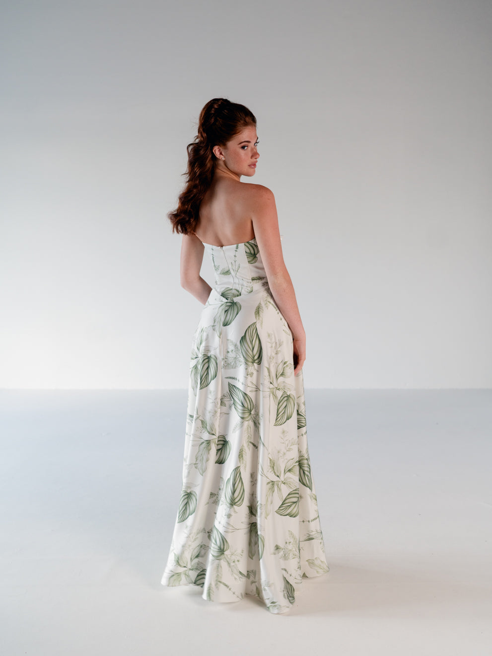 A woman stands in a studio wearing a strapless white maxi dress with a green leaf pattern, photographed from the back against a light gray background. / Basque waist draped bodice with an A-line skirt.