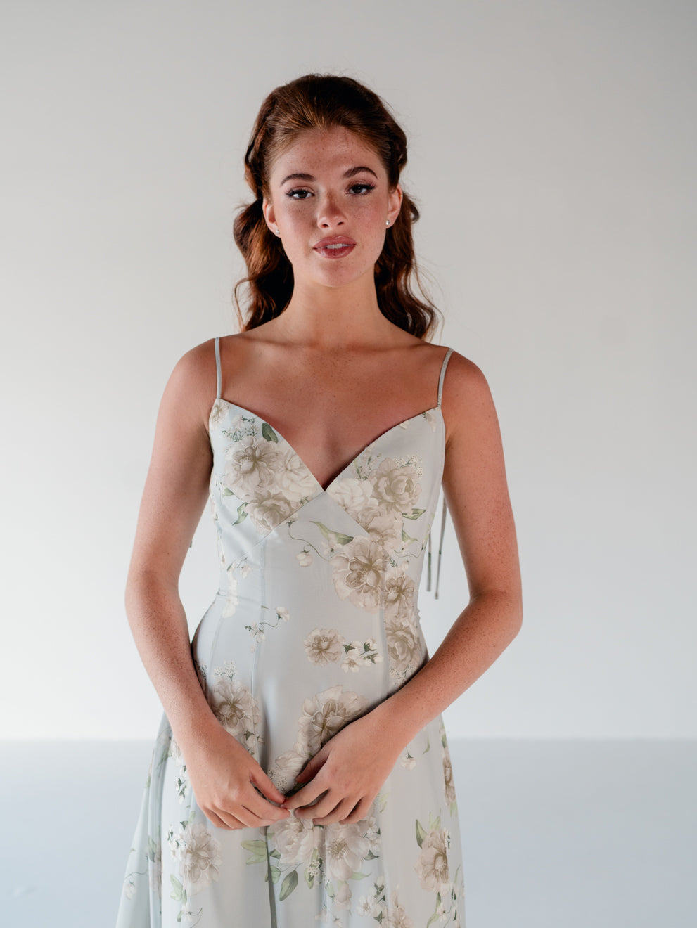 A woman modeling a light sage green midi dress with a beige and white floral print, featuring thin spaghetti straps and a sweetheart neckline, set against a plain light gray background. / A-line gown with double princess seam detail.